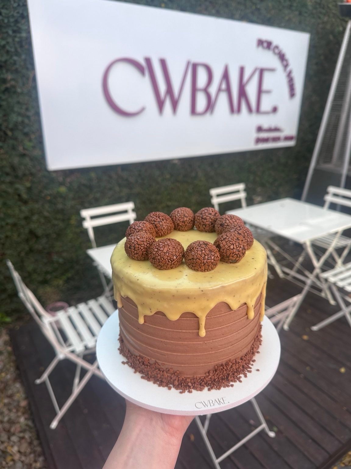 Bolo com drip de chocolate e brigadeiros no topo, em frente à fachada da CWBAKE