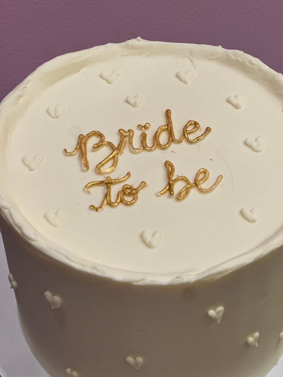 Bolo branco com a frase “Bride to be” em dourado e pequenos corações