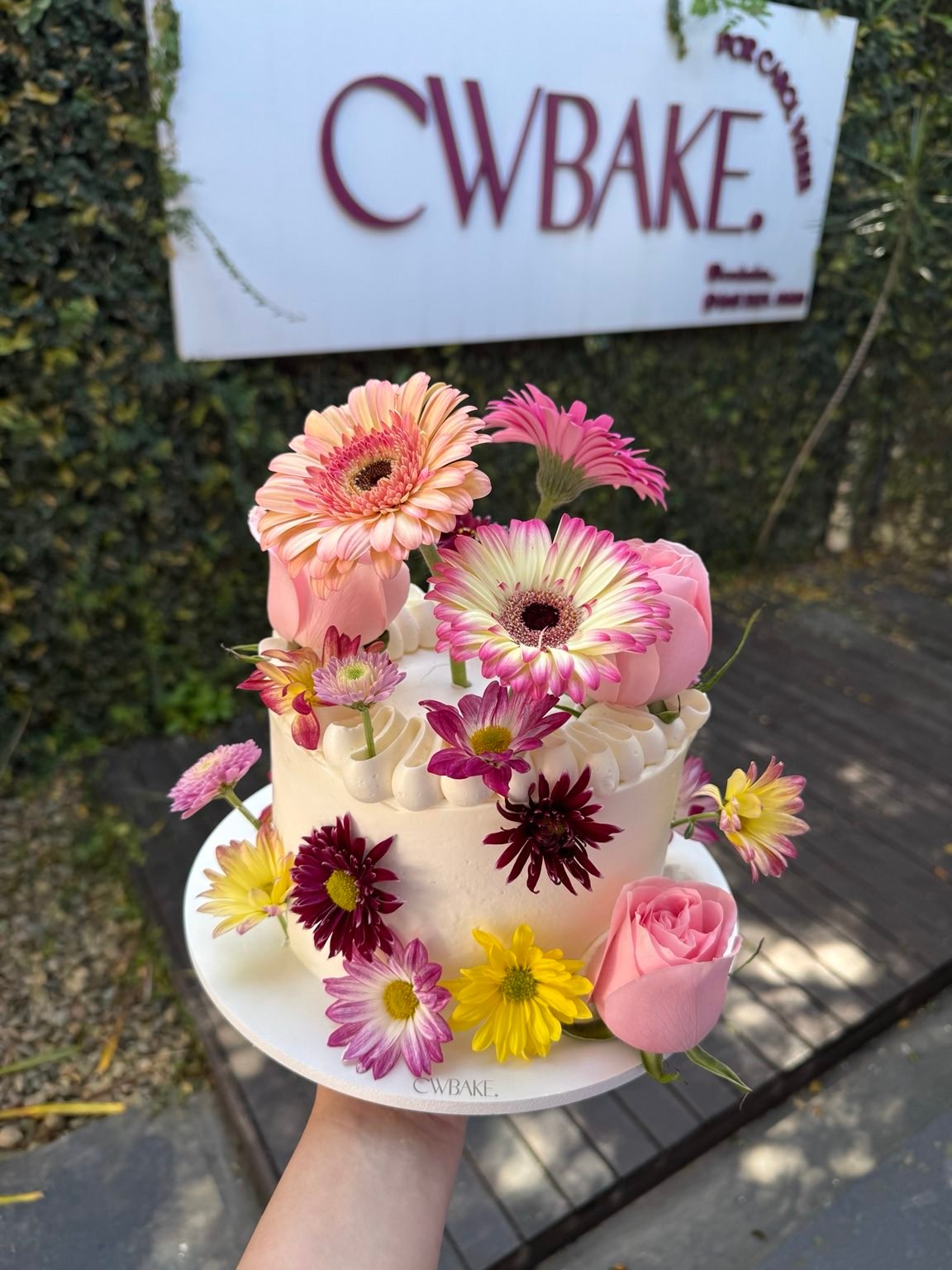Bolo com gérberas e flores naturais coloridas, em frente ao letreiro da CWBAKE